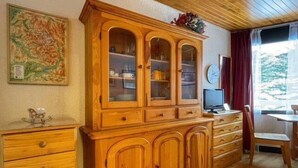 A wooden cabinet with multiple drawers and glass-fronted shelves, a small table with a chair, and a television on a stand.