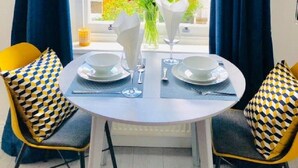 A dining area with a round table set for two, two chairs with patterned cushions, a vase with flowers, and a view of a brick building through a window with blue curtains. - Stunning studio with funky decor in Highgate village - perfect for couples! (London)