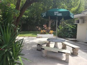 Un pati amb una taula de pedra i bancs, un parasol amb la inscripció "CAFE LA LLAVE ESPRESSO" i una font.