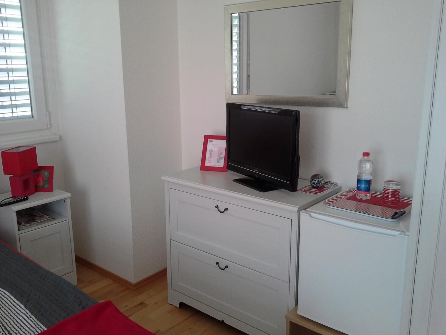 A bedroom with a bed, a white dresser with drawers, a television on top, a mirror above the dresser, and a window with blinds.