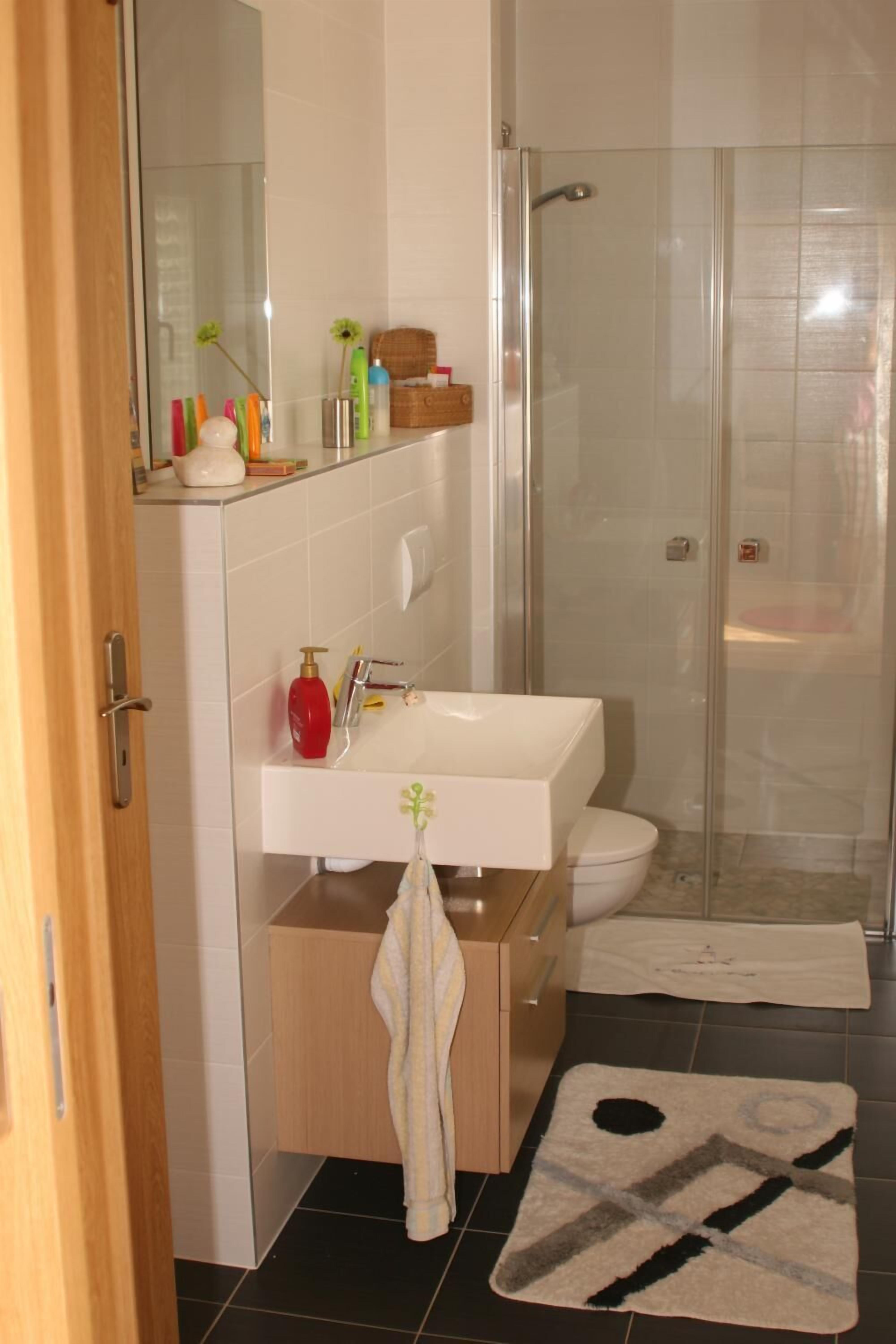 Une salle de bain avec une douche à parois de verre, un lavabo blanc et des toilettes. Une serviette est suspendue à un porte-serviettes, et des articles de toilette sont visibles sur le comptoir.
