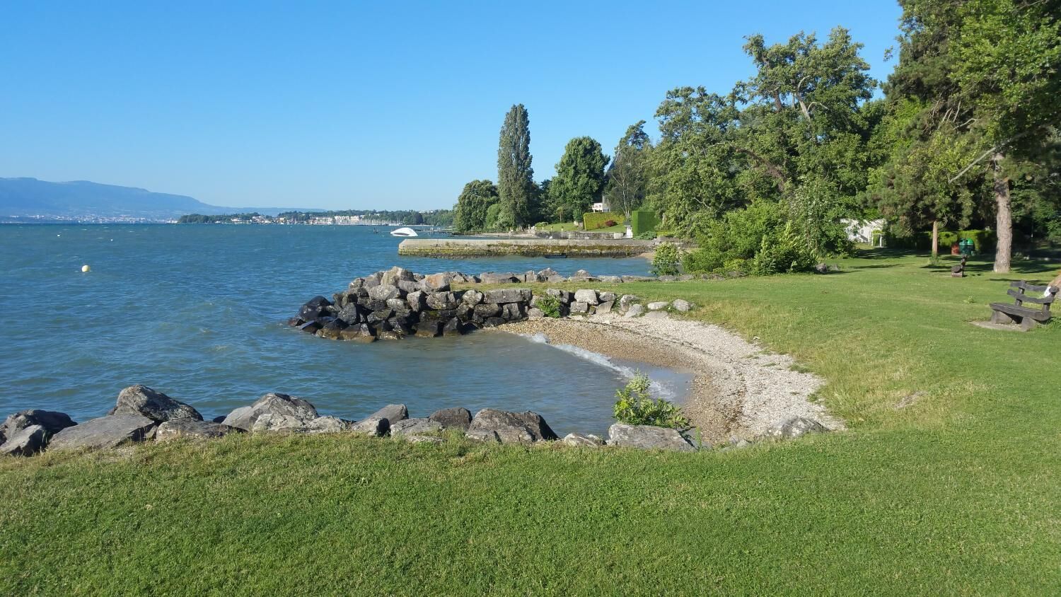 Un parc au bord du lac, avec une zone herbeuse, une rive rocheuse et une vue lointaine sur la silhouette d’une ville.