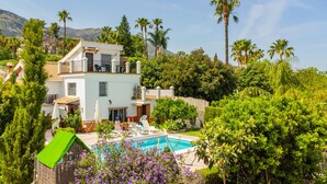 Una casa de dos pisos con piscina, rodeada de exuberante vegetación y palmeras.