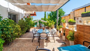 Un patio al aire libre con una mesa redonda y sillas, rodeado de plantas en macetas y una pérgola que ofrece sombra.