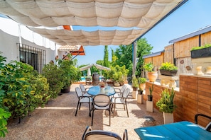 Een terras in de open lucht met een ronde tafel en stoelen, omgeven door plantenpotten en een pergola die schaduw biedt.