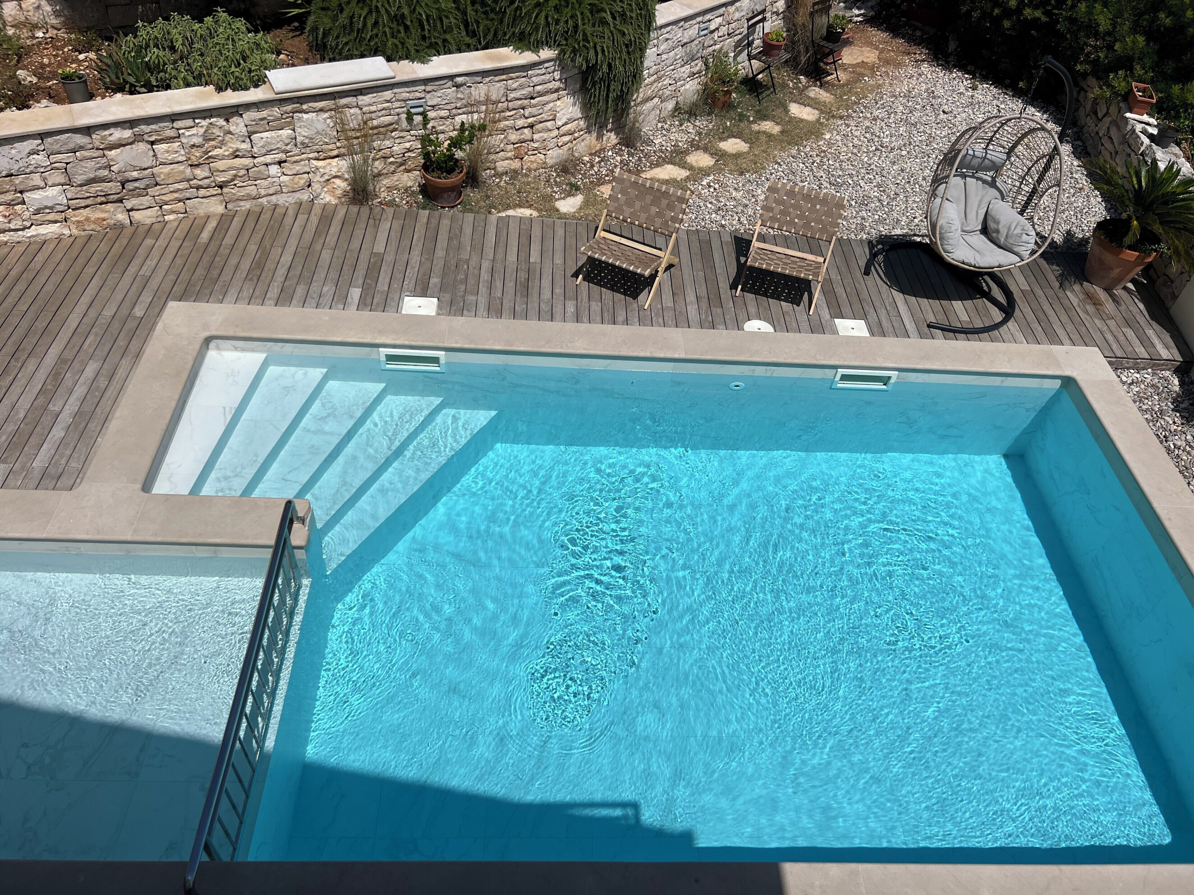 A rectangular swimming pool with a wooden deck surrounding it, stone walls, and outdoor seating.