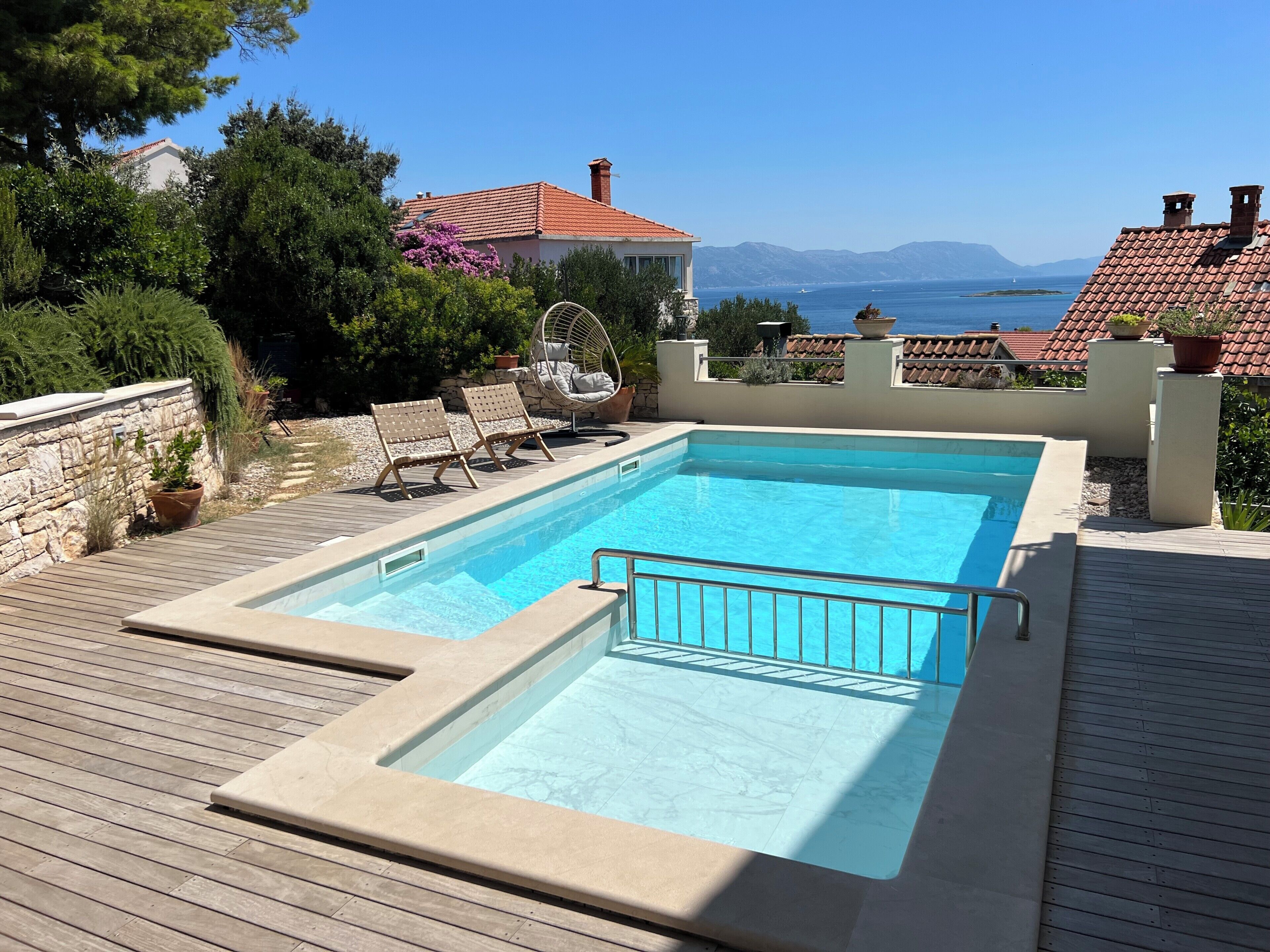 A rectangular swimming pool with a wooden deck, surrounded by stone walls and potted plants.