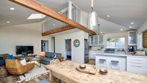 A spacious living area with a wooden dining table, a sofa, and a television. A kitchen with a large window and a hanging light fixture is visible. - Oceanfront Beauty with views from every room (Stinson Beach)