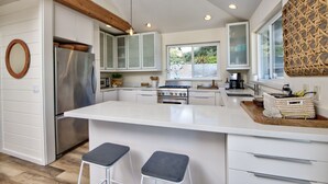 A modern kitchen with white cabinetry, a stainless steel refrigerator, a large island with stools, and a wooden ceiling beam. - Oceanfront Beauty with views from every room (Stinson Beach)