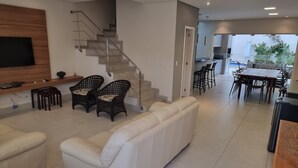 Ein modernes Wohnzimmer mit einer Couch, zwei Korbstühlen, einem an einer Holzvertäfelung montierten Fernseher, einer Treppe und einem Essbereich mit Tisch und Stühlen.