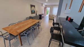Uma área de refeições com mesa e cadeiras de madeira, uma cozinha com bancada preta e chaleira vermelha, e uma sala de estar com sofá bege e um quadro na parede.