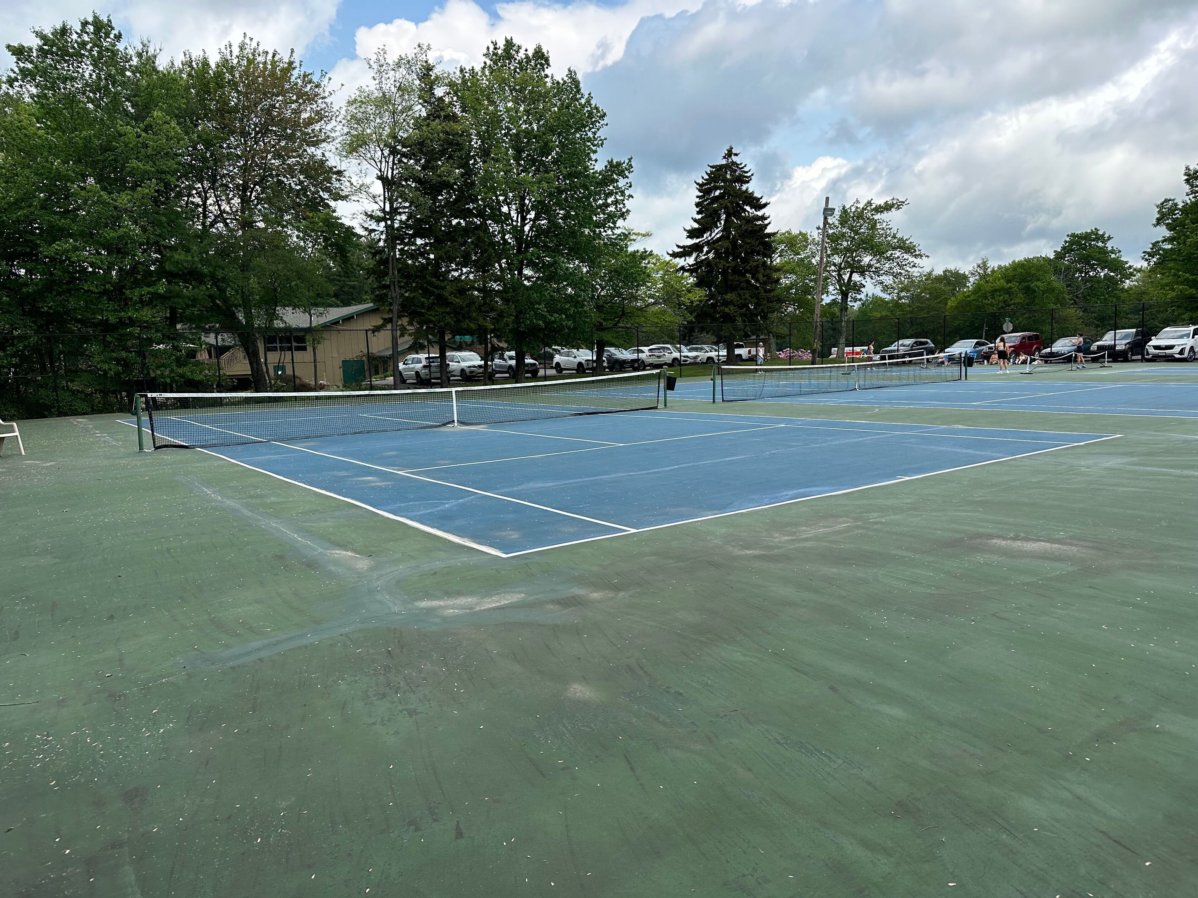 An outdoor tennis court with a net, surrounded by trees and parked cars.