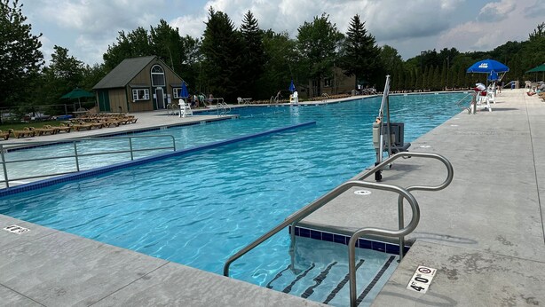 A swimming pool with clear blue water, surrounded by lounge chairs and umbrellas, with a building in the background.