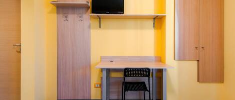 Une petite pièce avec un bureau en bois, une chaise noire, un téléviseur posé sur une étagère et une armoire.