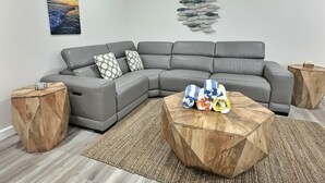 A modern living room with a grey sectional sofa, wooden side tables, a wooden coffee table, a rug, and wall art depicting a sunset and fish. - Beachside Vacation Rental Condo on Siesta Key with Pool Access (Siesta Key)