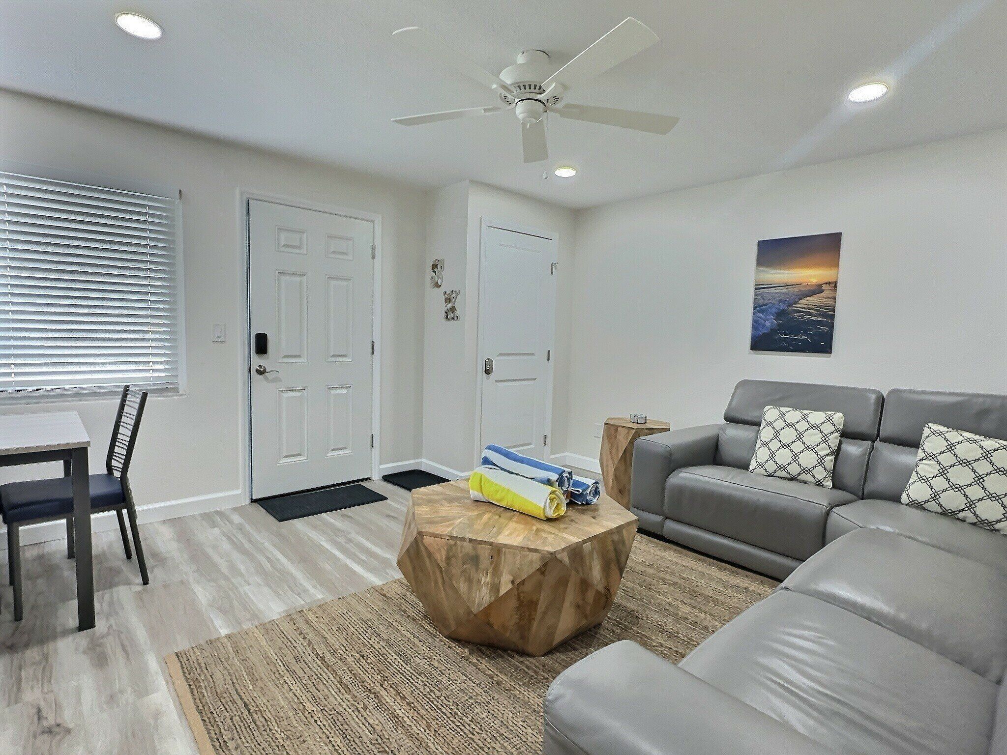A modern living room with a grey sofa, a wooden coffee table, a dining table with chairs, and a ceiling fan.