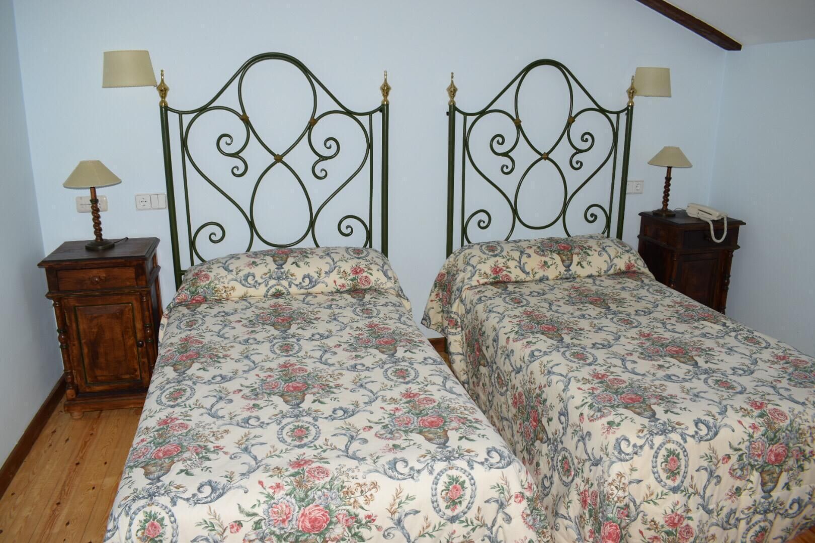 Una habitación con dos camas, cada una con cubrecama estampado con motivos florales, una mesita de noche de madera con lámpara y un pequeño aparador de madera también con lámpara.