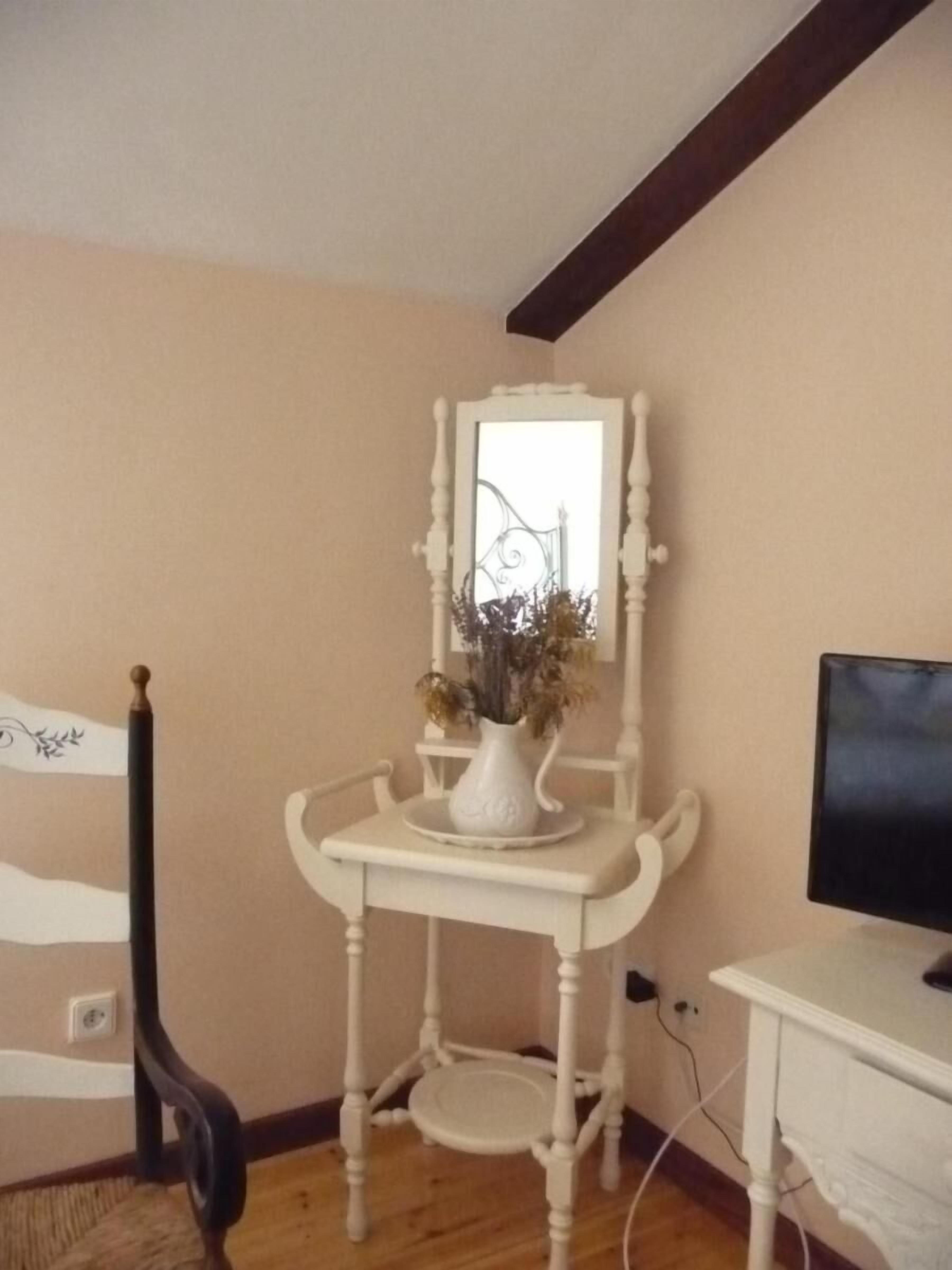 Une pièce avec une coiffeuse blanche, un miroir et un vase contenant des fleurs séchées.
