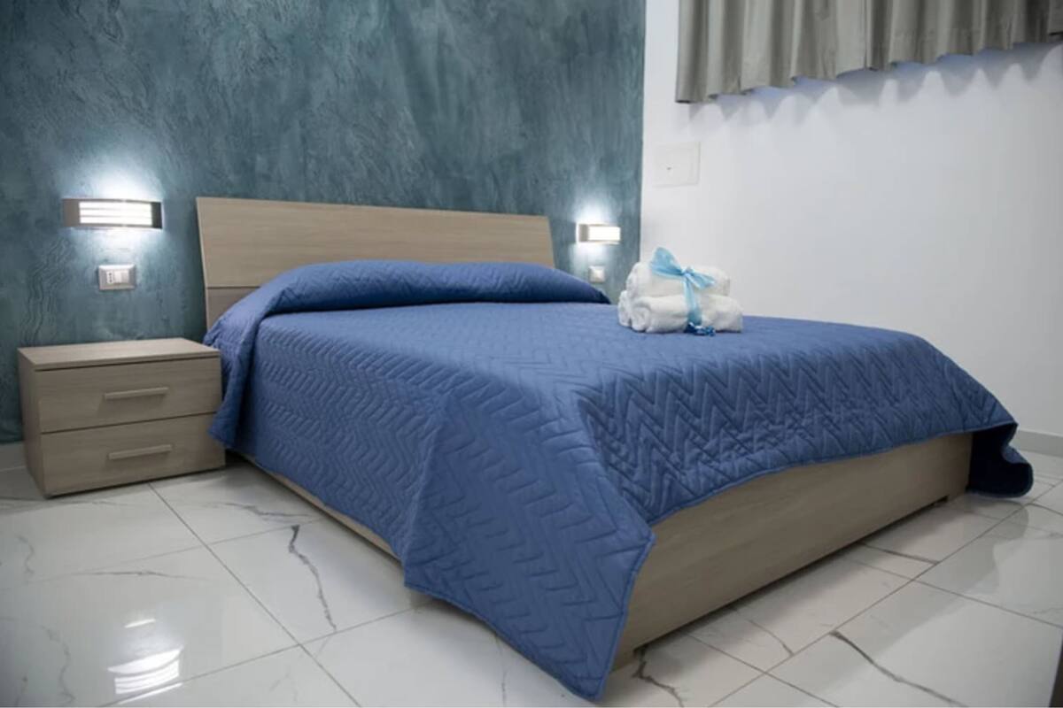 Un lit bien fait, recouvert d’une couette bleue, une table de chevet en bois avec des tiroirs et deux lampes fixées au mur.