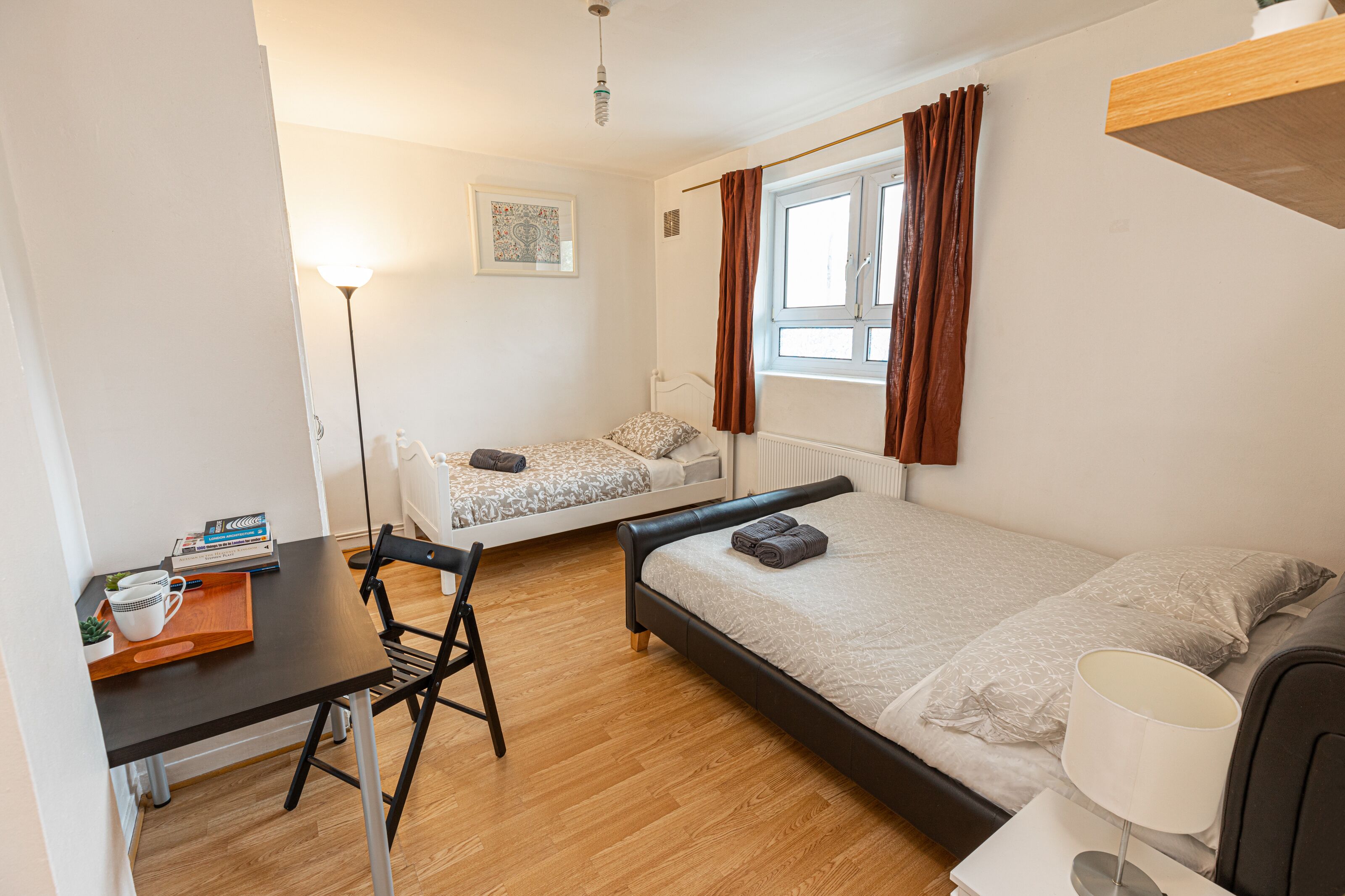 Et kompakt hotelv ærelse med en enkelt seng, et lille skrivebord med stol, et sengebord med lampe og et vindue med gardiner.
