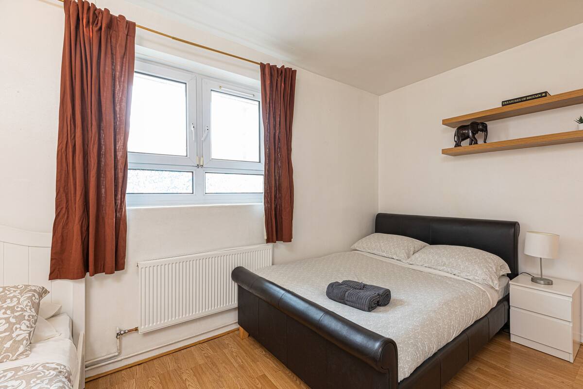 A bedroom with a bed, a window with curtains, a radiator, a bedside table with a lamp, and a shelf with a book and a small statue.