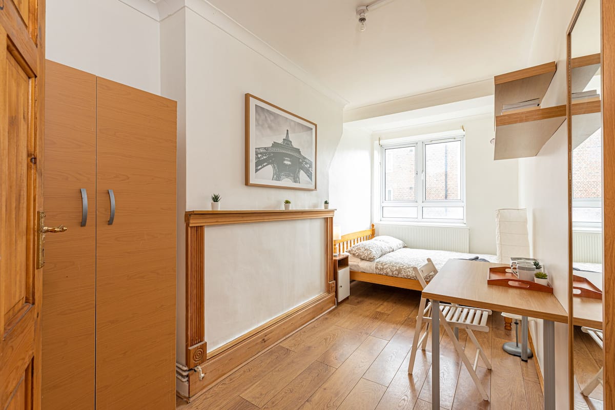 A bedroom with a bed, wooden wardrobe, a desk with a chair, a radiator, and a window.