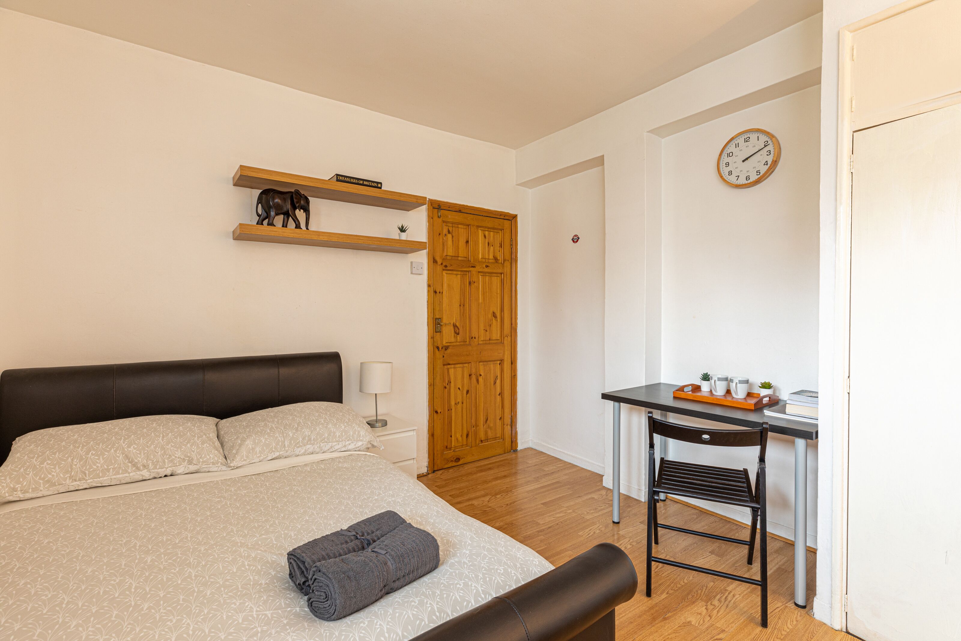 A bedroom with a bed, bedside table, lamp, wooden door, and a desk with a chair.