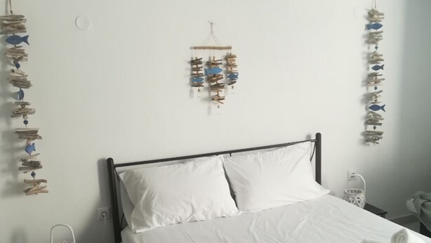 A neatly made bed with white linens and rolled towels, a bedside table with a lamp, and wall decorations including fish and wooden elements. - Classy Mykonian Studio - Ornos (Mykonos)