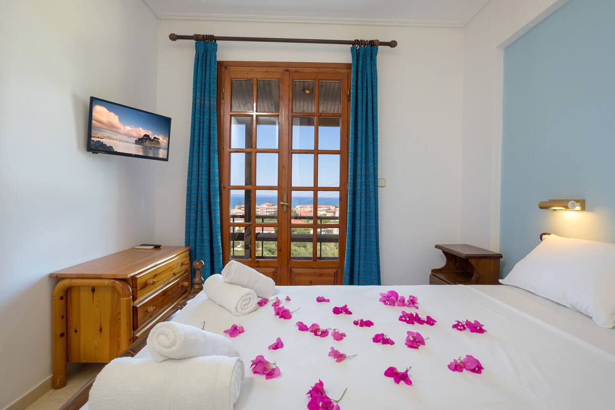 A bedroom with a bed, a wooden dresser, a nightstand, a wall-mounted TV, and a window with curtains.