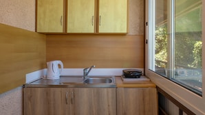 A compact kitchen with wooden cabinets, a stainless steel sink, a kettle, and a window providing a view of greenery outside. - Vallia's Apartments (Zakynthos)