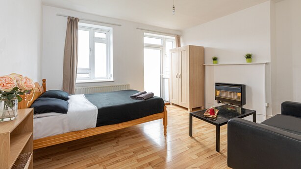 A bedroom with a bed, a wooden wardrobe, a television on a table, and a window with curtains. - Tower Bridge Accommodation - 5 (London)