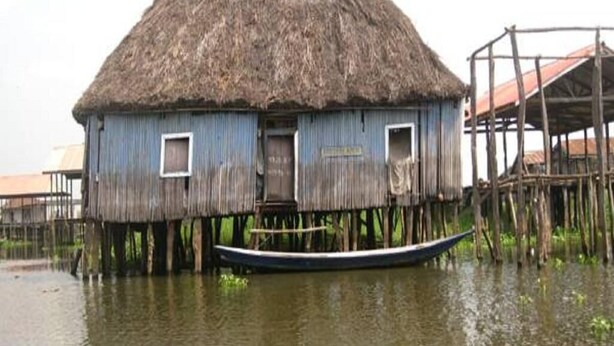 Uma casa de madeira tradicional com telhado de palha, erguida sobre estacas na água, com um barco atracado em frente.
