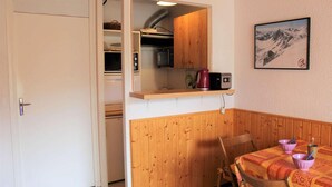 A compact kitchen with wooden cabinets, a microwave, and a kettle on a shelf. There is a dining area with a table set for two, featuring a patterned tablecloth and a painting of a mountain on the wall.