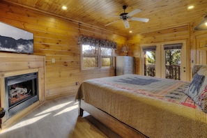 A cozy cabin room with a fireplace, a bed with a quilt, a ceiling fan, and a television on the wall. - Free Tickets | Great Views, Hot Tub, Fireplace, Jukebox, Theater & Arcade (Gatlinburg)