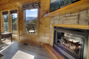 A wooden cabin interior with a stone fireplace, large windows, and a view of mountains. - Free Tickets | Great Views, Hot Tub, Fireplace, Jukebox, Theater & Arcade (Gatlinburg)