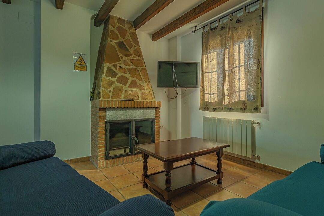 A living room with a stone fireplace, a television mounted on the wall, and a window with curtains.