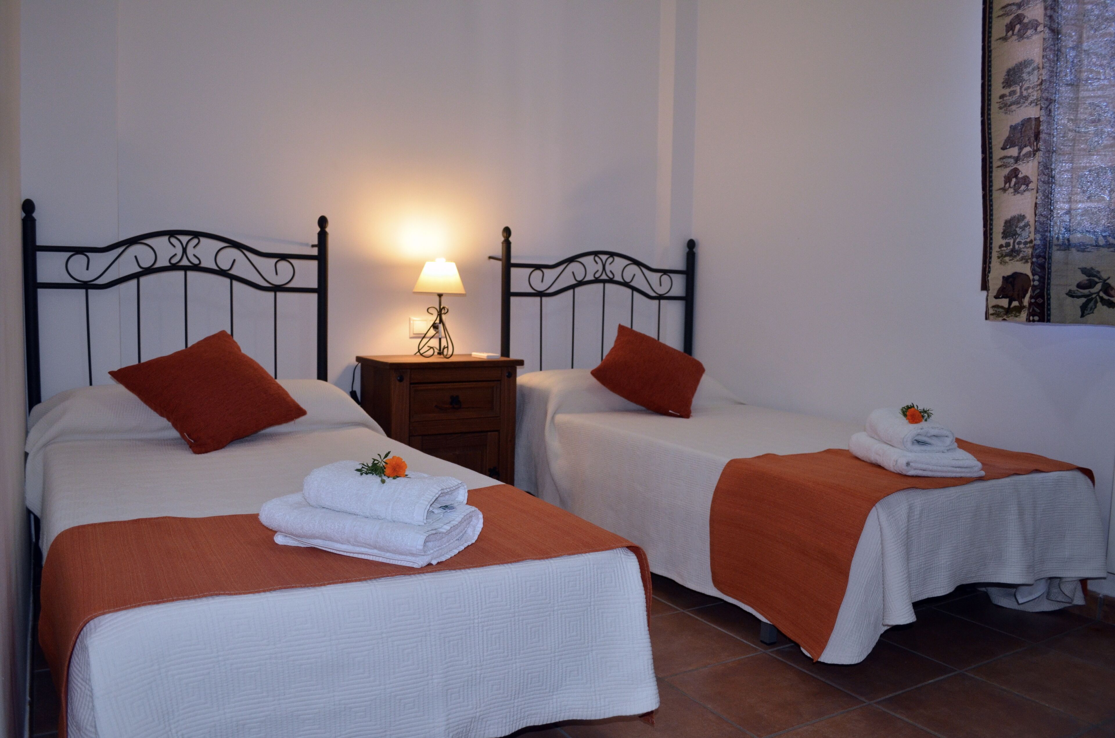 A hotel room with two single beds, each with an orange blanket and a white pillow. There is a nightstand with a lamp between the beds.