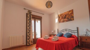 A bedroom with a red bedspread, two pillows, a wooden nightstand with a lamp, a radiator, a window with curtains, and a painting on the wall. - Casa Rural Mirador de los Tomillos (Cardena)