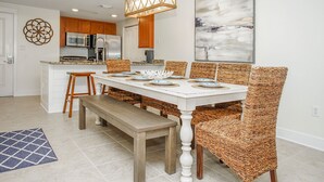 A dining area with a white table, wicker chairs, and a bench. There is a kitchen in the background with wooden cabinets and stainless steel appliances. - Waterscape B210 COMPLETE REMODEL OCEAN & POOL VIEW! BEACH SERVICE! 2 KINGS BEDS! (Fort Walton Beach)