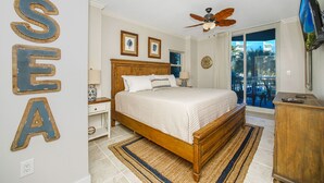 A bedroom with a wooden bed, two bedside tables, a ceiling fan, a large window, and a view of the outdoors. - Waterscape B210 COMPLETE REMODEL OCEAN & POOL VIEW! BEACH SERVICE! 2 KINGS BEDS! (Fort Walton Beach)