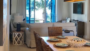 A dining area with a table set for four, a sofa, and a television in the background. - Waterscape B210 COMPLETE REMODEL OCEAN & POOL VIEW! BEACH SERVICE! 2 KINGS BEDS! (Fort Walton Beach)
