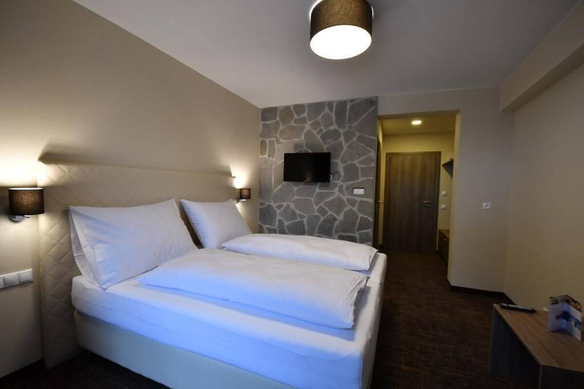 Habitación de hotel con una cama grande, un muro decorativo de piedra, un televisor de pantalla plana sobre la cama y una puerta de madera que conduce a otra habitación.
