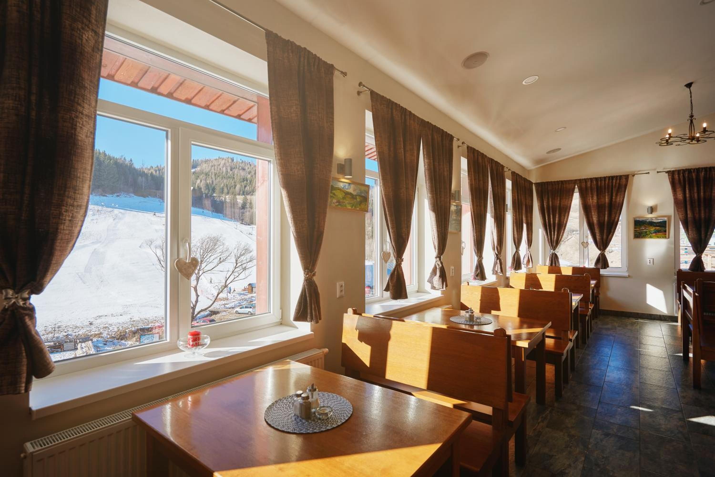 A restaurant with wooden tables and chairs, large windows with curtains, and a view of a snowy landscape.