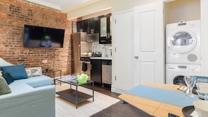 A compact living space with a brick wall, a flat-screen TV, a sofa, a coffee table, a dining table with chairs, a kitchen area with cabinets, a microwave, a stove, and a washing machine. - Apartments Nadim (Skopje)