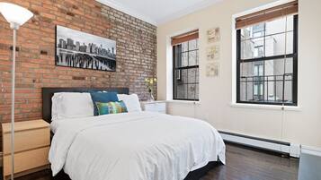 A bedroom with a brick wall, a bed with white bedding, a nightstand, a floor lamp, and two windows with blinds.