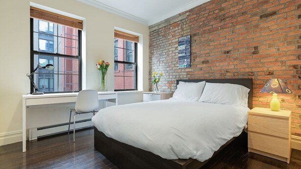 A bedroom with a large bed, a desk, and a chair. There are two windows with blinds, a brick wall, and a wall-mounted sign. - Apartments Nadim (Skopje)