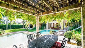 Um espaço ao ar livre coberto com piscina, mesa e cadeiras, e vista para uma casa ao fundo.