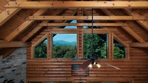 A log cabin with a sloped roof, large windows offering mountain views, a ceiling fan, and a rustic interior design. - Peaceful Paradise secluded mountain log cabin with scenic view and game room (Whittier)