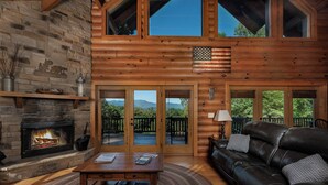 A log cabin interior with a fireplace, leather sofa, and a view of mountains through large glass doors. - Peaceful Paradise secluded mountain log cabin with scenic view and game room (Whittier)