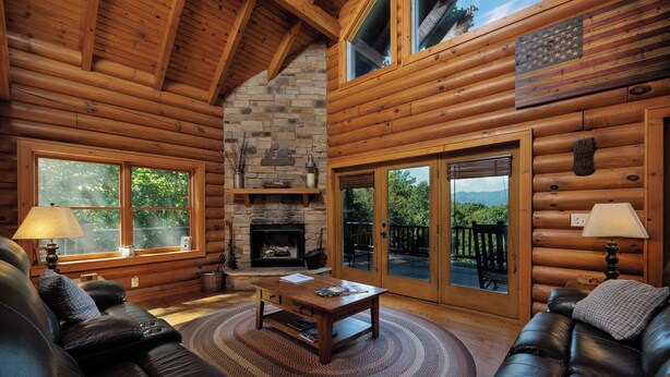 A log cabin interior with a fireplace, leather sofas, a coffee table, and large windows offering a view of the outdoors. - Peaceful Paradise secluded mountain log cabin with scenic view and game room (Whittier)